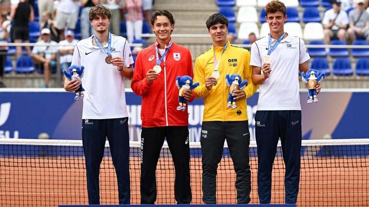 Four men wearing, from left to right, white, red, yellow and white sports tops, all showing of medals hung around their necks. A tennis net is directly behind them, and crowds are visible in stands in the background