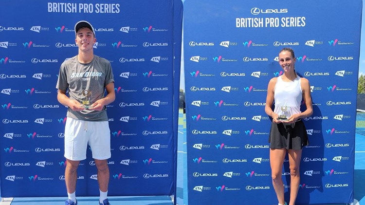 Two people, one man and one woman, stood holding glass trophies in front of a large blue board with small sponsor logos on it