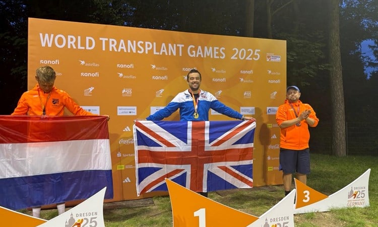 A man holding a British union flag stands in front of a large orange backdrop displaying the words 'World Transplant Games 2025'