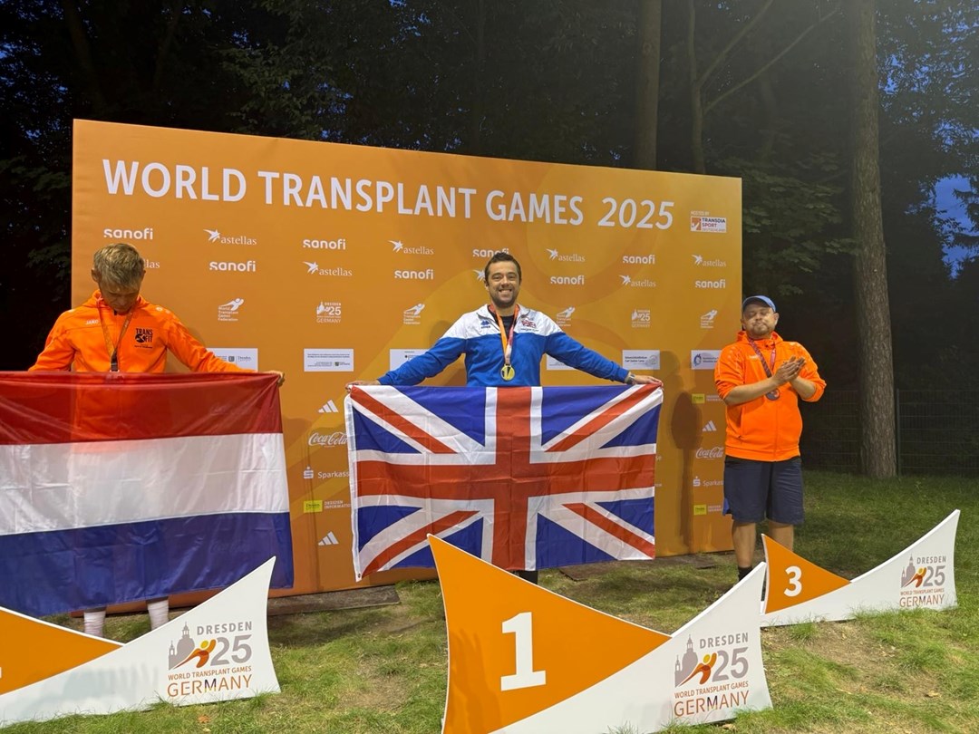 A man holding a British union flag stands in front of a large orange backdrop displaying the words 'World Transplant Games 2025'