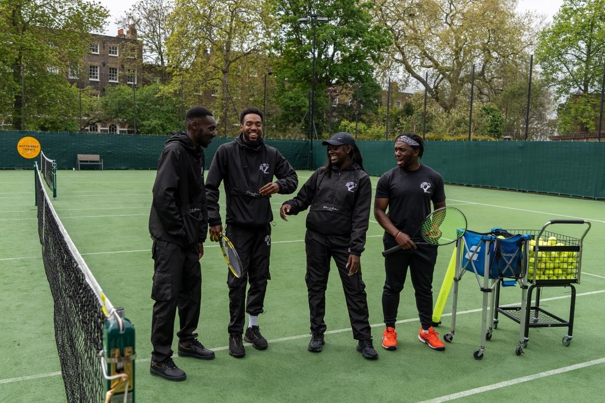 A group of four black people, all wearing black clothing, are stood together laughing on a tennis court, holding racquets and standing near a ball cart filled with tennis balls.