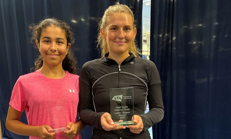 A girl in black short and a black t-shirt smailes at the camera, holding a glass trophy engraved with the words, 'Deaf Tennis National Finals Ladies' Singles Winner 2025'. Next to her stanbds a girl wearing a pink t-shirt, also holding a glass trophy and smiling