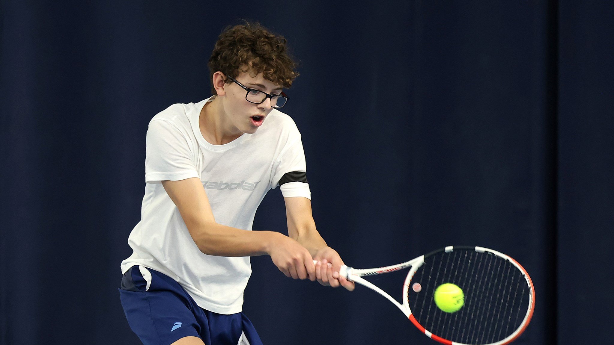 A boy wearing a white t-shirt, blue shorts and glasses hits a tennis balls. His mouth is slightly open with exertion