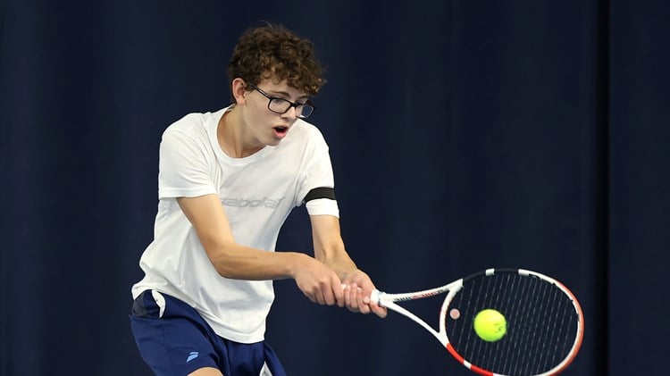 A boy wearing a white t-shirt, blue shorts and glasses hits a tennis balls. His mouth is slightly open with exertion