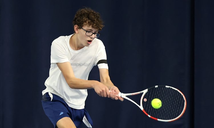 A boy wearing a white t-shirt, blue shorts and glasses hits a tennis balls. His mouth is slightly open with exertion