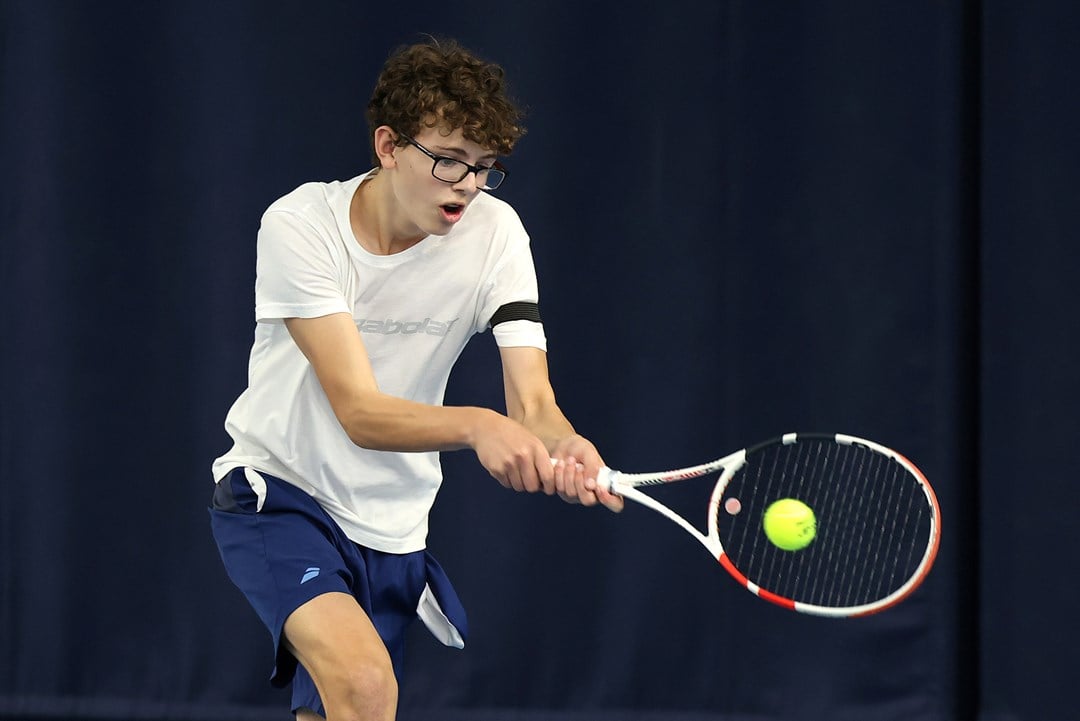 A boy wearing a white t-shirt, blue shorts and glasses hits a tennis balls. His mouth is slightly open with exertion