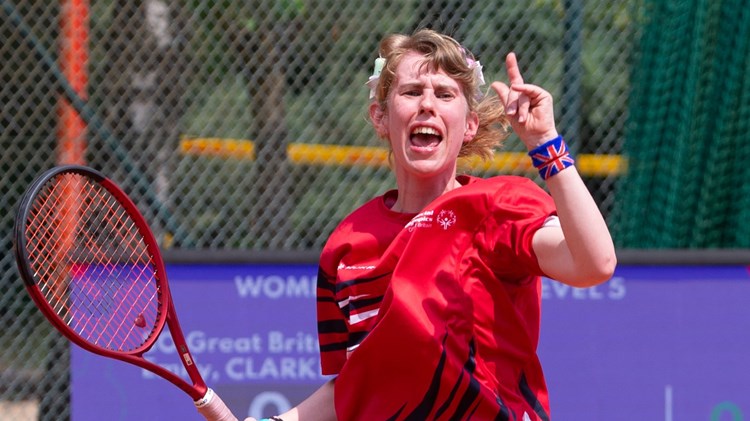 A girl in a red sports t-shirt celebrates, and holds a tennis racket in her right hand. On her left wrist, she is wearing a wristband with the British union flag on it.