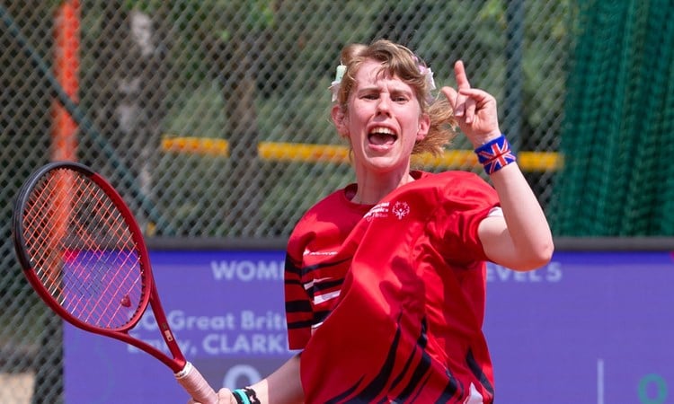 A girl in a red sports t-shirt celebrates, and holds a tennis racket in her right hand. On her left wrist, she is wearing a wristband with the British union flag on it.