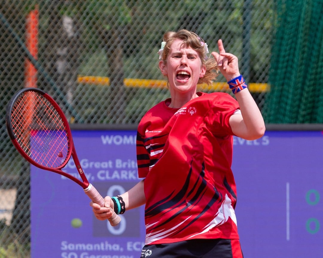 A girl in a red sports t-shirt celebrates, and holds a tennis racket in her right hand. On her left wrist, she is wearing a wristband with the British union flag on it.