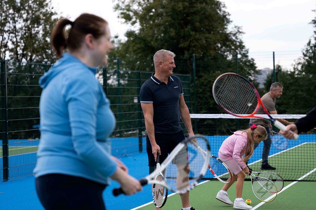 Sports Minister Stuart Andrew playing tennis alongside a family at refurbished tennis courts