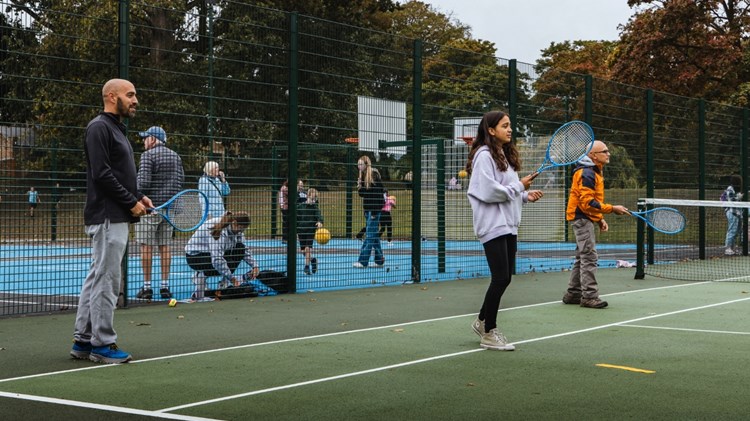 Three peopl two adult men and a young girl, each hold blue tennis racets in theire hands, while standing on a green tennis court. There is fencing behind them, spearting the tennis court from an area where adults and children are bouncing balls, and basketball hoops can be seen