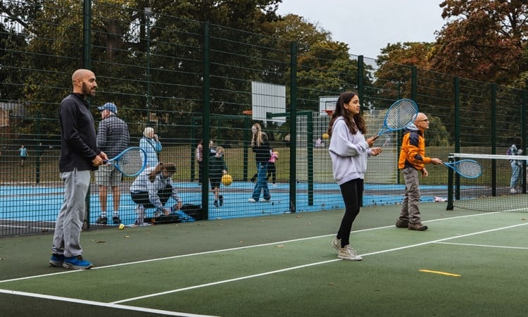 Three peopl two adult men and a young girl, each hold blue tennis racets in theire hands, while standing on a green tennis court. There is fencing behind them, spearting the tennis court from an area where adults and children are bouncing balls, and basketball hoops can be seen