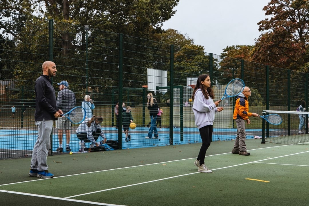 Three peopl two adult men and a young girl, each hold blue tennis racets in theire hands, while standing on a green tennis court. There is fencing behind them, spearting the tennis court from an area where adults and children are bouncing balls, and basketball hoops can be seen