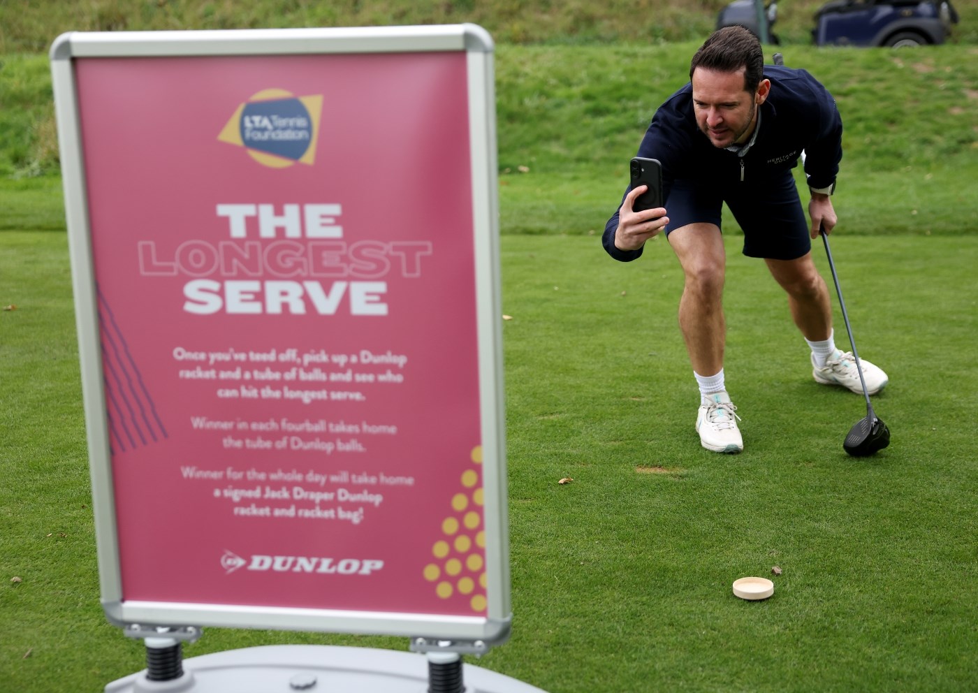 A man wearing dak shorts and a dark shirt holding a golf club bends down holding a phone in his right hand to take a photograph of a large pink sign. The sign has the words 'The Longest Serve' written in large font, along with smaller writing underneath