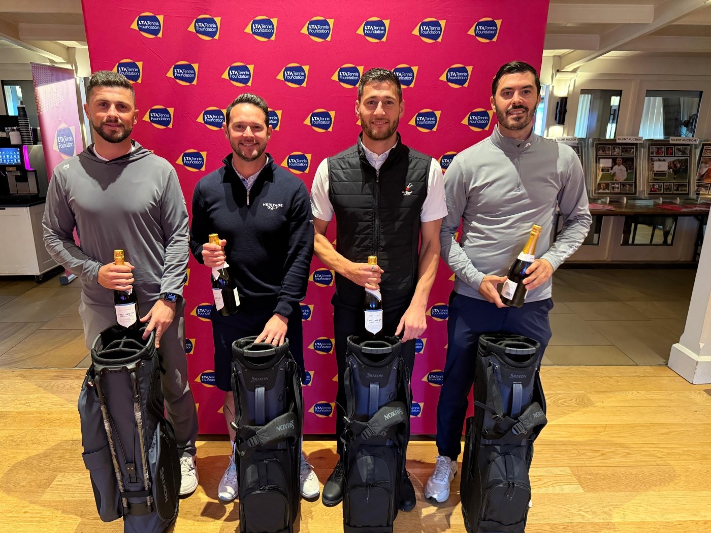 Four men stand in front of a large pink backdrop, each holding a bottle of champagne and each supporting a large black golf bag.