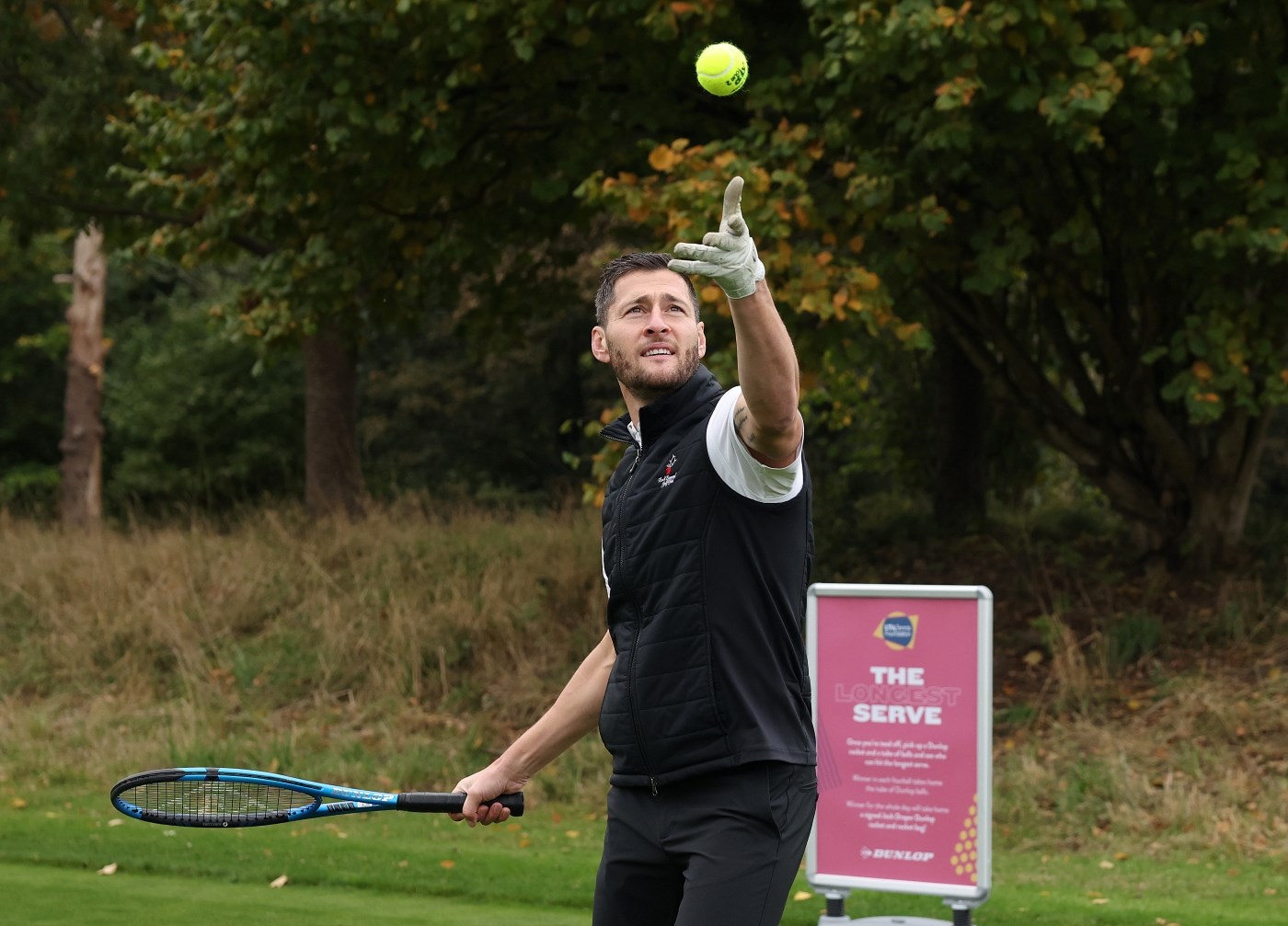 A man prepares to hit a tennis ball that he has thrown into the air with a blue tennis racket. Behind him, green grass and trees are visible, along with a sign on which the words, 'The Longest Serve' are visible.