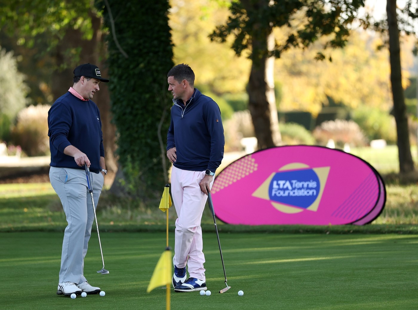 Two men, both wearing dark blue shirts and grey trousers, stand with golf balls at their feet and each holding a putter in their hand. They stand on a large area of grass, and behind them , a lare banner with the words, 'LTA tennis Foundation' is visible.