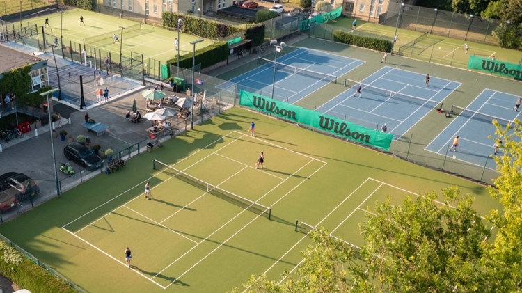 A wide view of green tennis courts with people playing on them, with one court in the foreground obscured by foliage from a large tree. In the background, there a a number of blocks of flats, in the bottom left corner, there is a white car parked behind a hedge. 