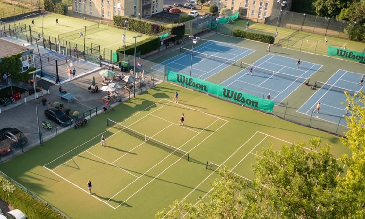 A wide view of green tennis courts with people playing on them, with one court in the foreground obscured by foliage from a large tree. In the background, there a a number of blocks of flats, in the bottom left corner, there is a white car parked behind a hedge. 