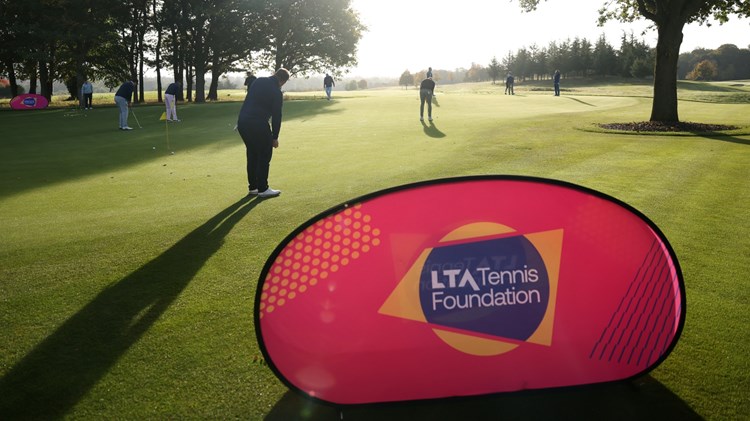 A large pink banner with the words 'LTA Tennis Foundation' is positioned on a large area of grass. Behind the banner, a group of people is spread out around a large flat area of grass, with many of hem holding small golf clubs. The sun is shining bright, with trees seen stretching off into the distance.