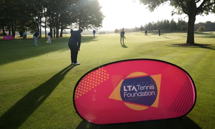 A large pink banner with the words 'LTA Tennis Foundation' is positioned on a large area of grass. Behind the banner, a group of people is spread out around a large flat area of grass, with many of hem holding small golf clubs. The sun is shining bright, with trees seen stretching off into the distance.