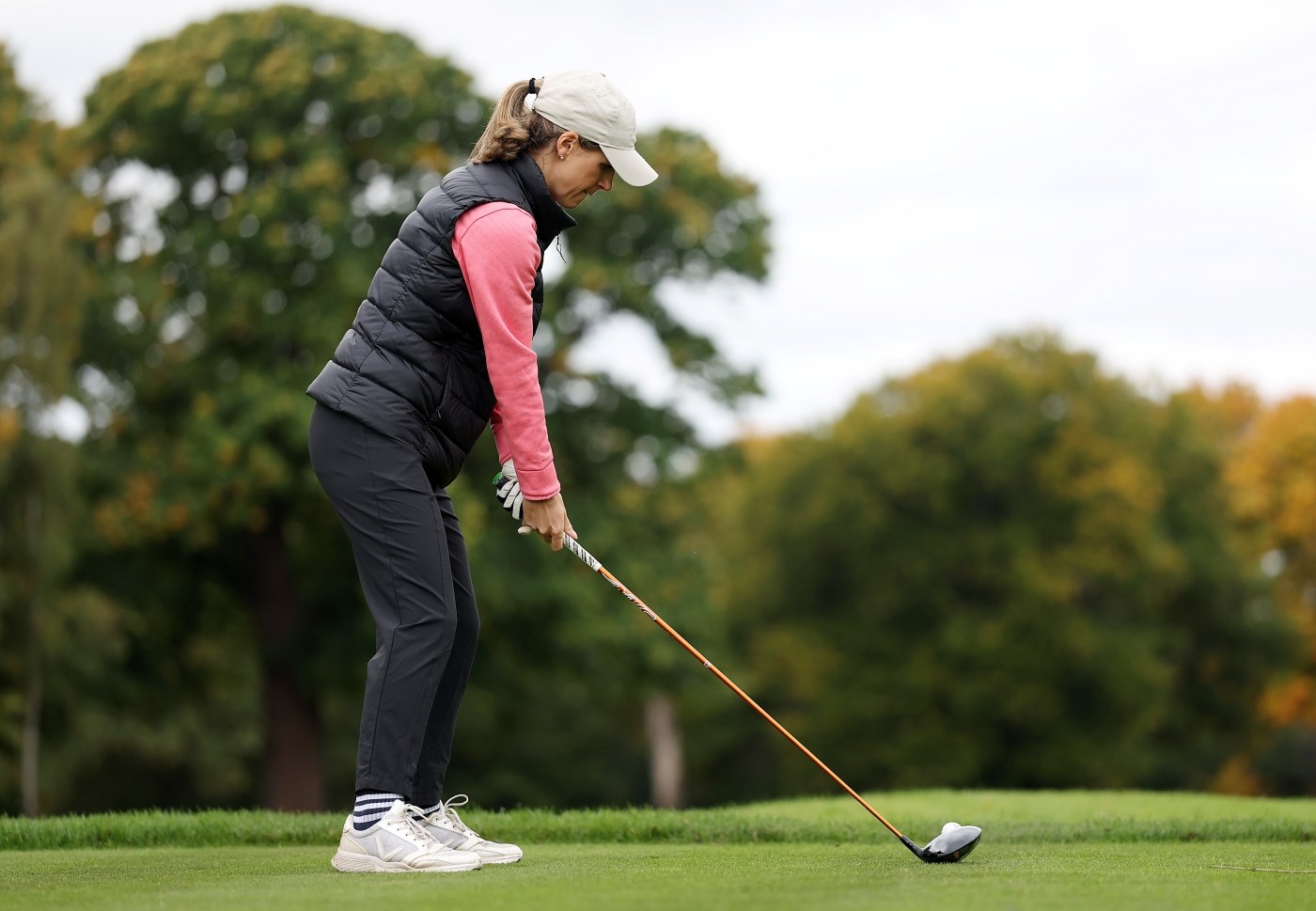 A woman wearing black trousers, and black gilet and a pink shirt prepares to hit a golf ball with a large golf club. Large trees with a variety of green and yellow leaves are out of focus in the background.