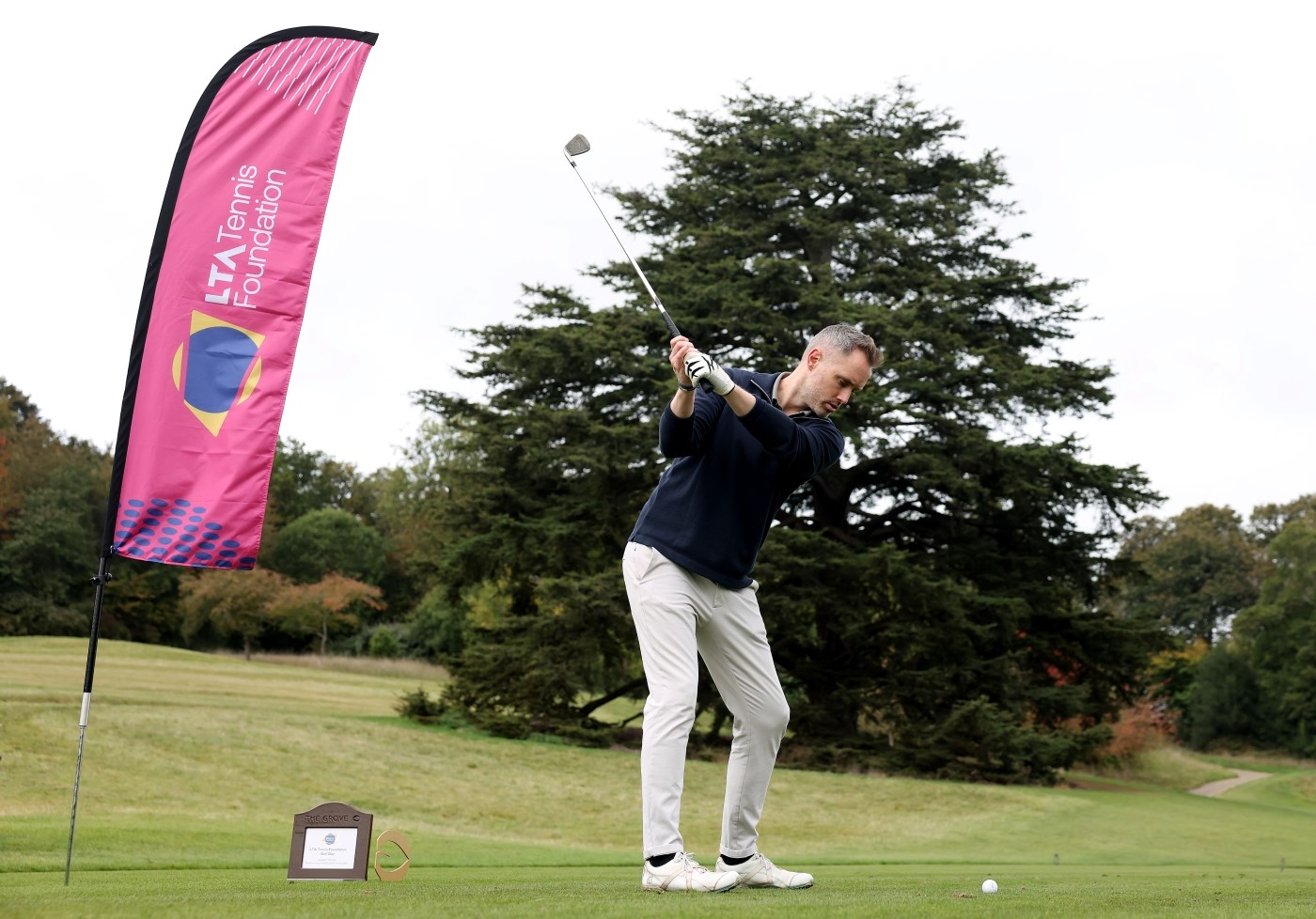 A man wearing a dark blue long-sleeved top and white trousers raises his golf club about to hit a golf ball. To his left is a large pink flag with the words, 'LTA tennis Foundation' on it. Trees and open green space up a hill are visible behind the man.