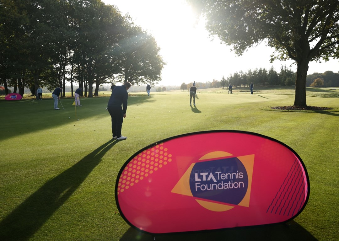 A large pink banner with the words 'LTA Tennis Foundation' is positioned on a large area of grass. Behind the banner, a group of people are spread out around a large flat area of grass, with many of them holding small golf clubs. The sun is shining bright, with trees seen stretching off into the distance.