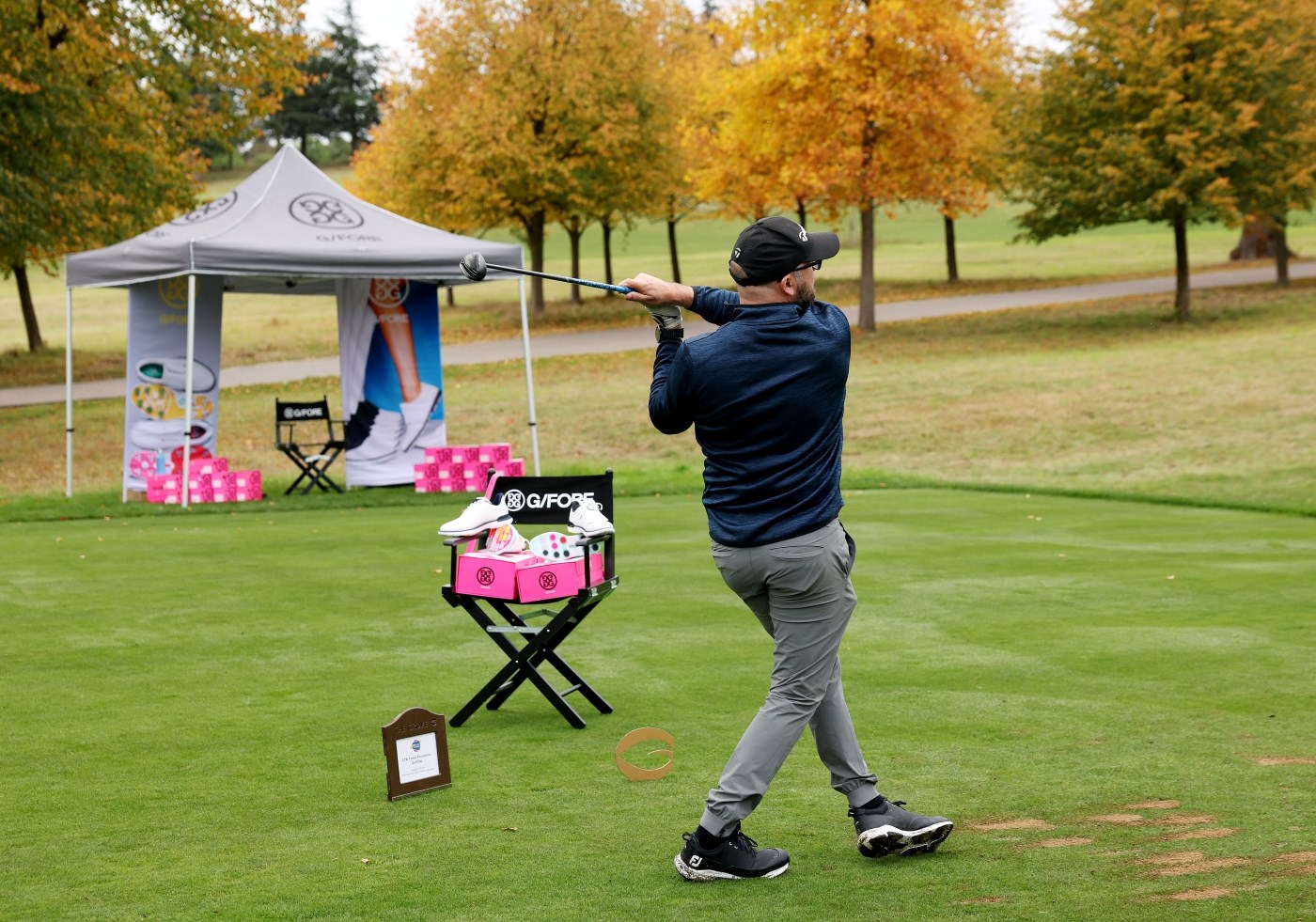 A man wearing a dark blue shirt and grey trousers holds his stance after hitting a golf shot. To this left, a chair is positioned with white shoes and pink shoeboxes placed on top. Beyond the chair, a gazebo with a grey roof is set up, and more pink shoeboxes can be seen. Trees with green and yellow leaves can be seen in the background.
