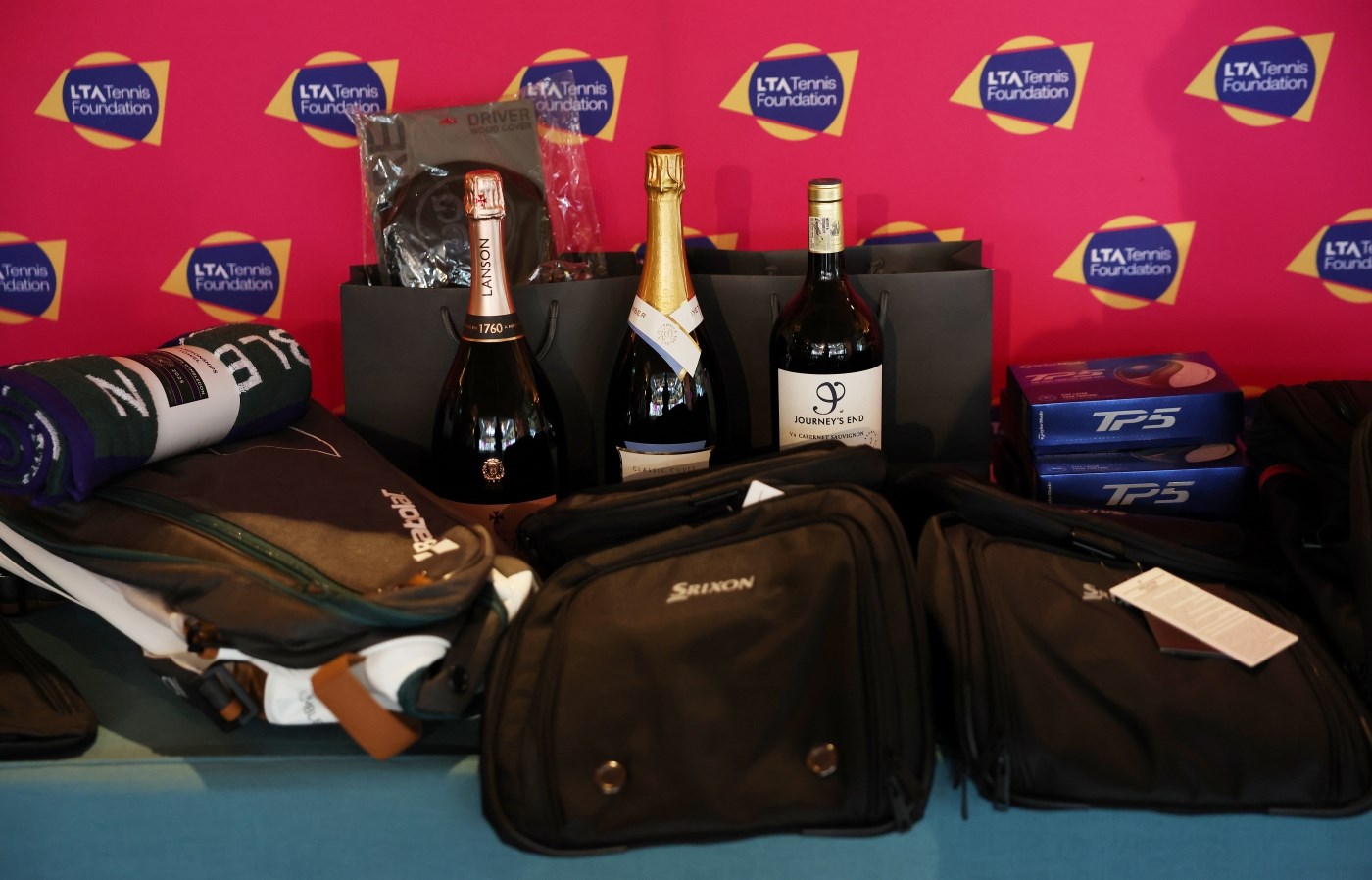 A selection of bags, a green towel, three bottles of wine in a row in the middle and two blue boxes to the right of the wine all sit in front of a pink backdrop, with repeated logos featuring the words, 'LTA Tennis Foundation'.