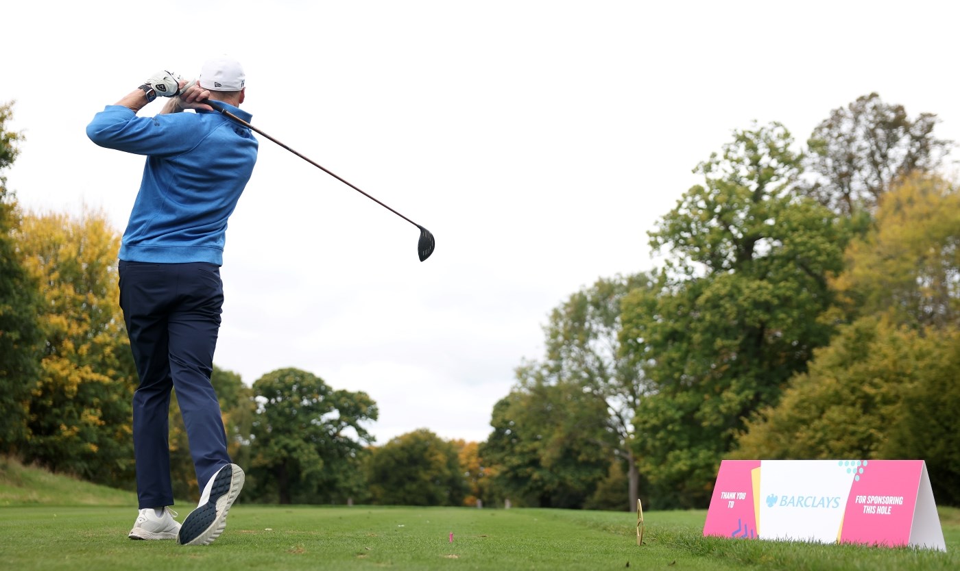 A player wearing dark blue trousers, and a light blue long-sleeved top and a white cap stands tall holding a golf club, having just hit a golf ball into the distance. Trees are visible in the distance, and to the person's right, a pink and white triangular prism with the word 'Barclays' in blue font on a white background is visible.