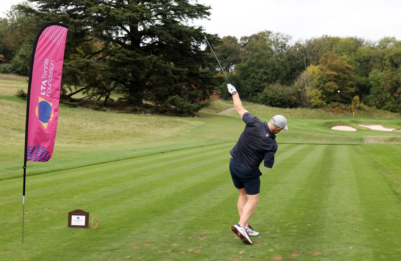A man wearing a black shirt and black shorts, who is missing his right arm, hits a golf ball towards a yellow flag that is visible in the distance. To his left, a large flag with the words, 'LTA Tennis Foundation' is planted in the ground. Trees are visible to the player's left and far in the distance.