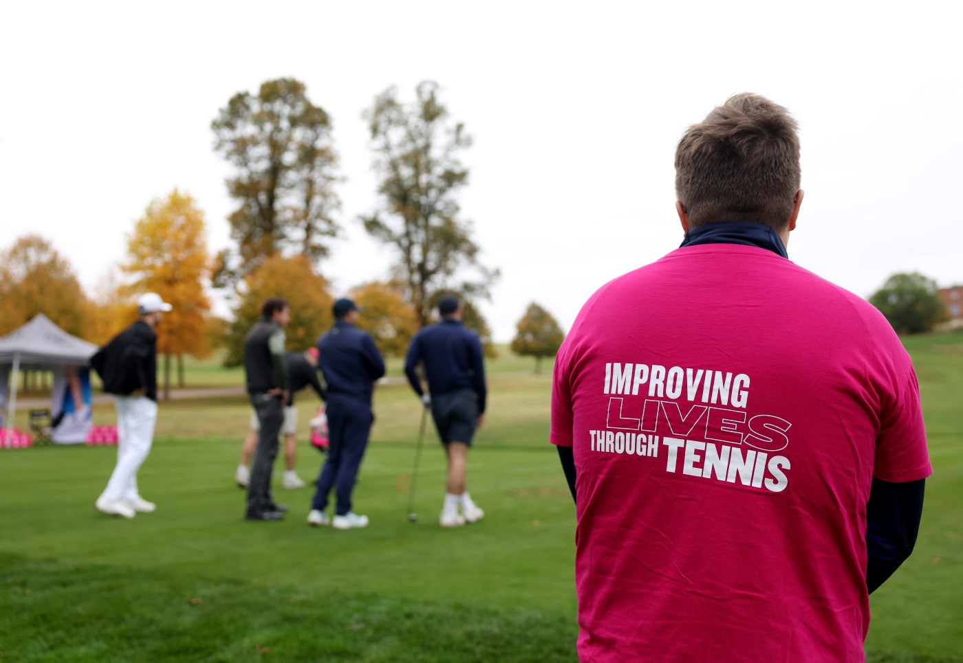 A person stands looking at a group of golfers in the distance. Their back is facing the camera, and they are wearing a pink t-shirt with the words, 'Improving Lives Through tennis' written on the shirt.