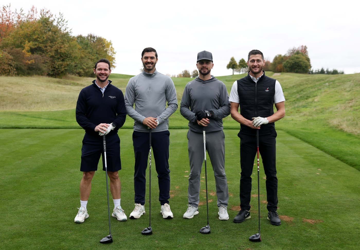 Four men stand smiling and holding golf clubs on a large patch of grass. Behind them to their right, a large patch of trees is visible, with more trees stretching further into the distance.