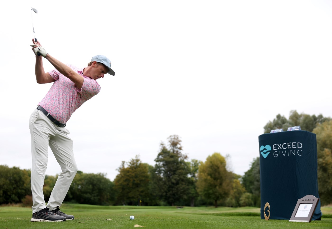 A man wearing a pink shirt, white trousers and a white hat holds his golf club in the air about to swing and hit a golf ball. To the man's right, a blue pedestal with the words, 'Exceed Giving' is positioned, whilst trees can be seen in the distance.