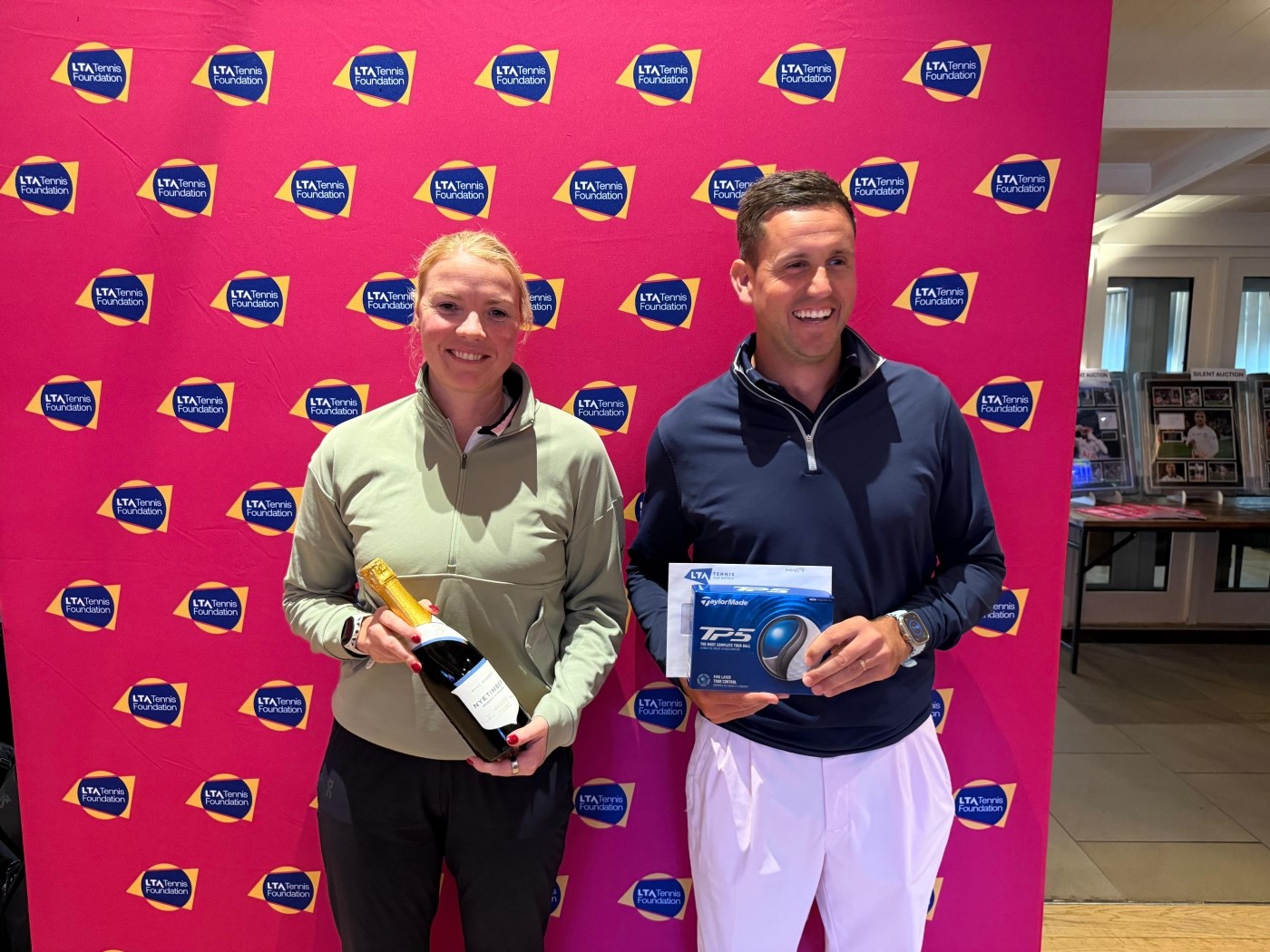 A woman wearing a light green shirt stands smiling, holding a bottle of sparkling wine. Next to her, a man wearing a dark blue top and white trousers is smiling, holding a box of golf balls. Both are standing in front of a large pink backdrop.