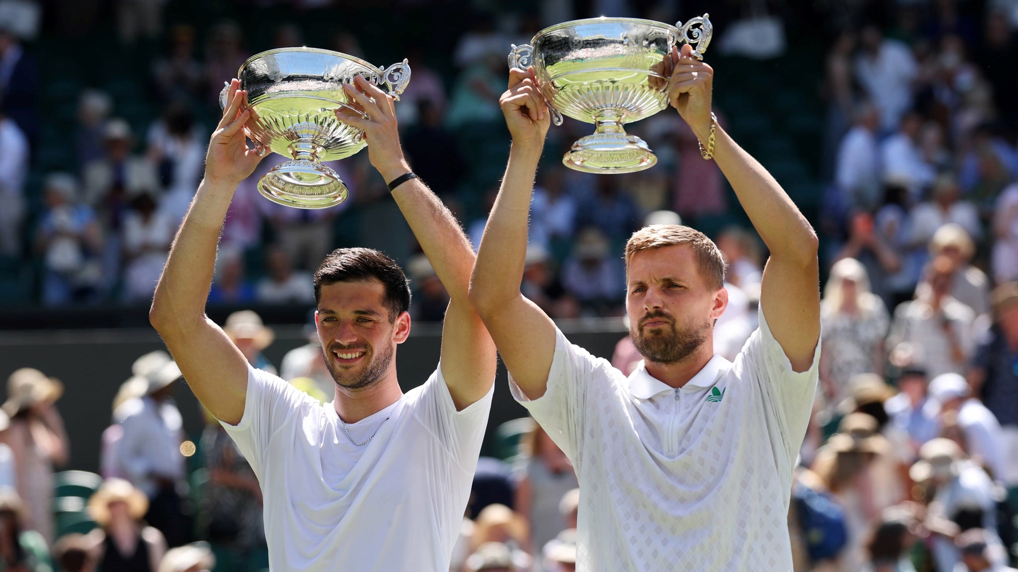 Julian Cash and Lloyd Glasspool smiling while holding up the Wimbledon men's doubles trophies above their head in front of the crowd