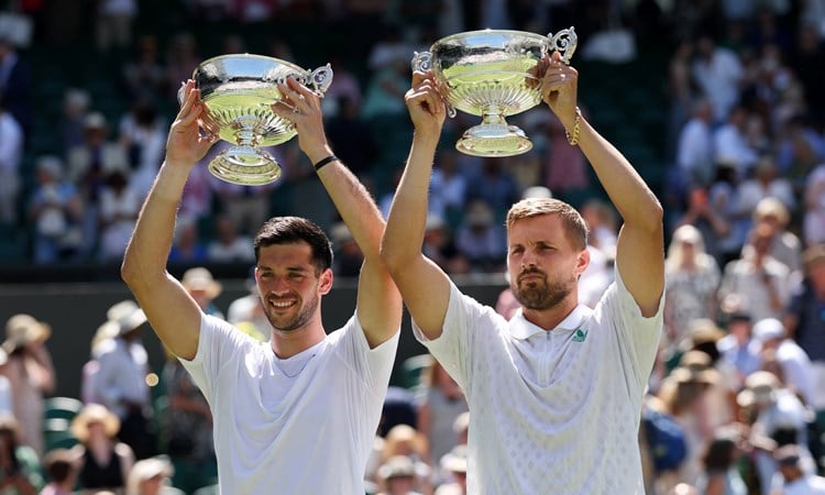 Julian Cash and Lloyd Glasspool smiling while holding up the Wimbledon men's doubles trophies above their head in front of the crowd