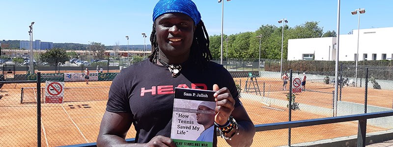 Sam Jalloh holding his book in front of tennis courts