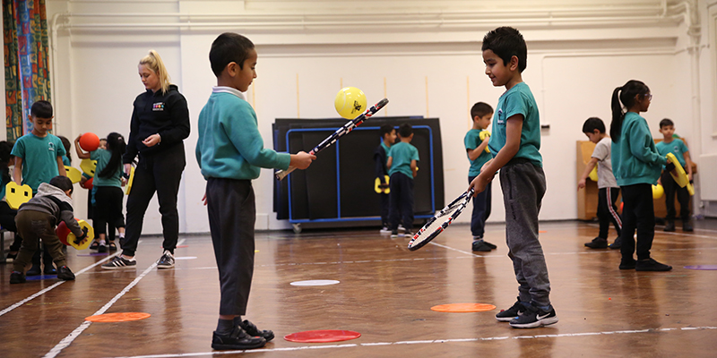 Two children in their school hall hitting a ball to each other with tennis rackets
