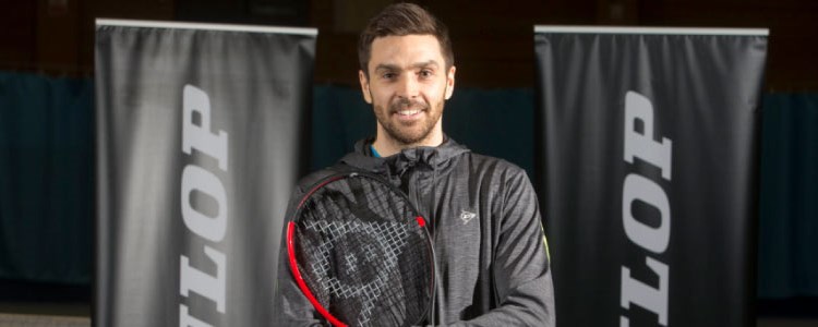 colin fleming  smiling for a picture  wearing all back with a racket in hand and dunlop  banners at the back