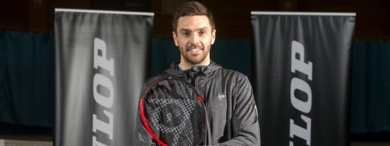 colin fleming smiling for a picture wearing all back with a racket in hand and dunlop banners at the back