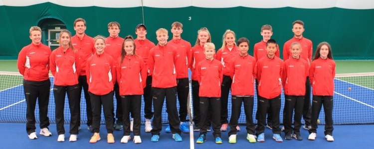 Group picture of the junior team wales standing on court in front of the net wearing red jackets and smiling