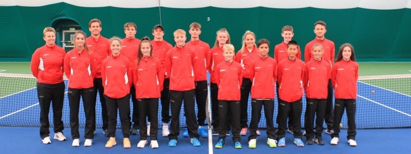 Group picture of the junior team wales standing on court in front of the net wearing red jackets and smiling