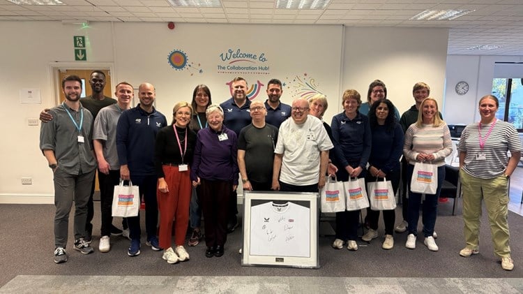 A group of people standing together looking at the camera for a group photo. In the middle is a framed tennis shirt signed by British players 