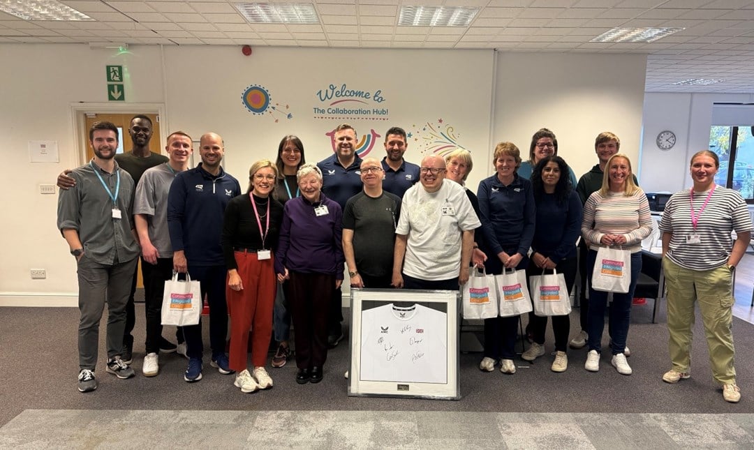 A group of people standing together looking at the camera for a group photo. In the middle is a framed tennis shirt signed by British players