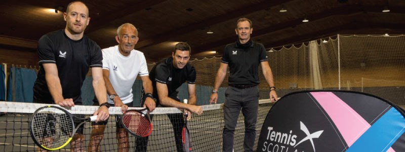 four tennis players standing and leaning on the net with rackets in hand next to a tennis scotland  banner