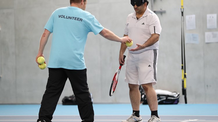 Volunteer hands a tennis ball to a visually impaired player