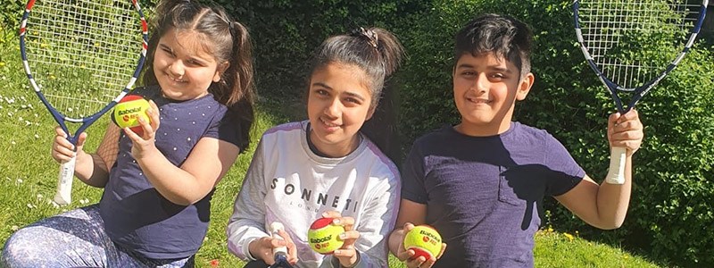 three kids smiling all holding a yellow and red tennis ball and two with tennis rackets in the air