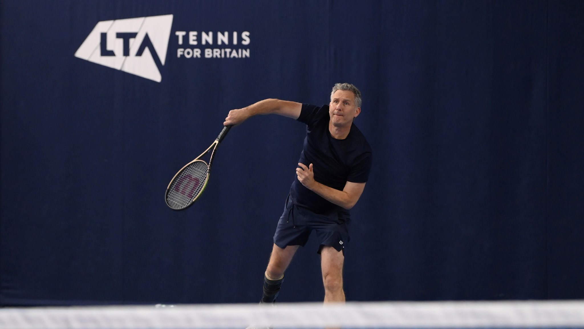 Comedian and tv presenter Adam Hills serving on court while playing para standing tennis at the National Tennis Centre in Roehampton